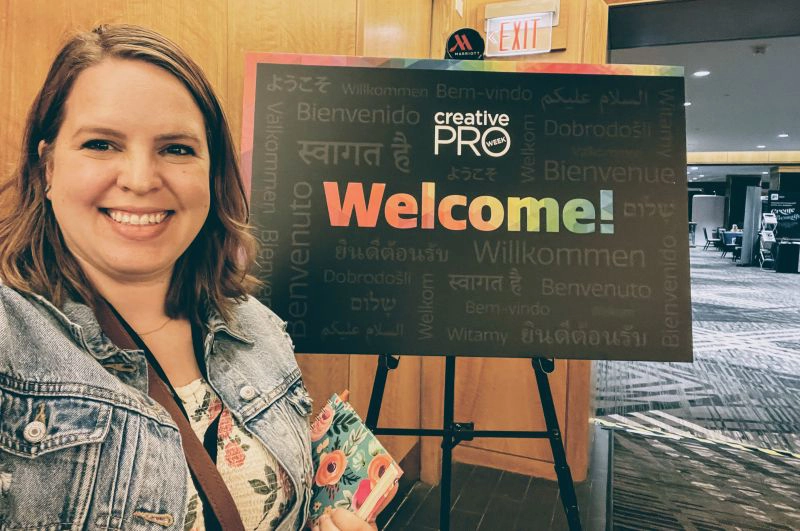 Lindsey smiles broadly at a conference center in front of a CreativePro Week welcome sign.